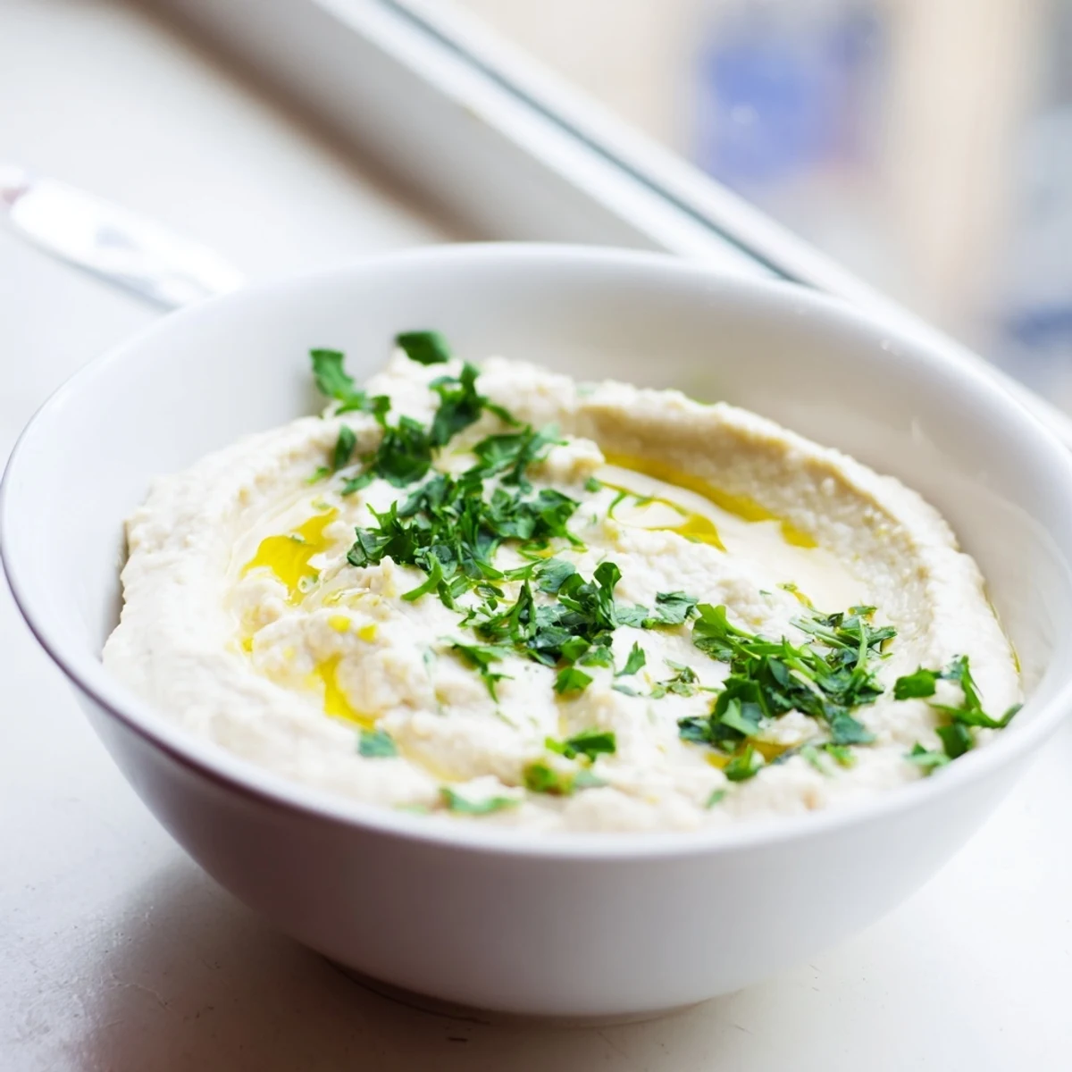 A bowl of creamy White Bean Dip topped with a generous drizzle of olive oil and fresh chopped parsley, served with pita chips.