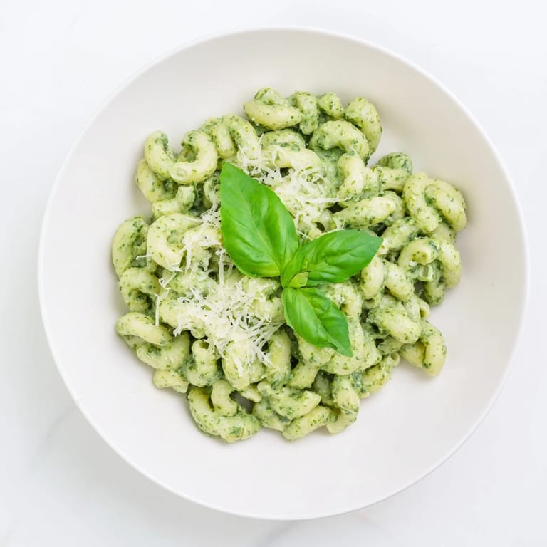 A close-up of Creamy Avocado Pesto Mac with creamy avocado sauce, ripe avocado slices, and fresh basil leaves on a sunny table setting.